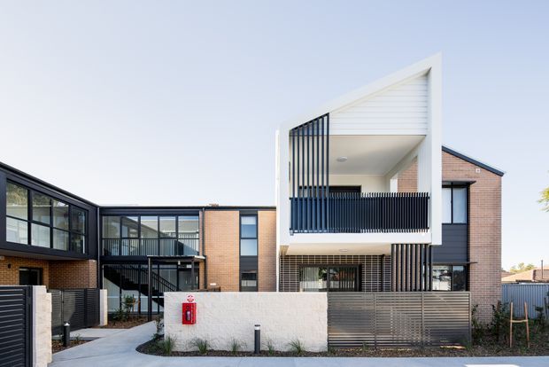 Street-facing view of a contemporary multi-storey residential development with several attached brick and dark-clad dwellings. The prominent dwelling features a projecting upper-level balcony, white geometric gable-like framing, vertical dark privacy screens, large glazed openings, and a rendered boundary wall with horizontal metal screening. A shared paved access path and neighbouring attached units are visible.