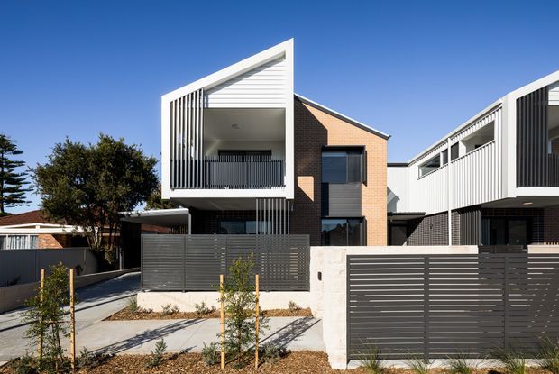 Street-facing view of a contemporary multi-unit residential development with sharply framed projecting balconies, white vertical screens and balustrades, dark horizontal metal fencing, exposed red-brick walls, and concrete paving. The buildings have asymmetric pitched roof forms and a clean, geometric composition in bright daylight.