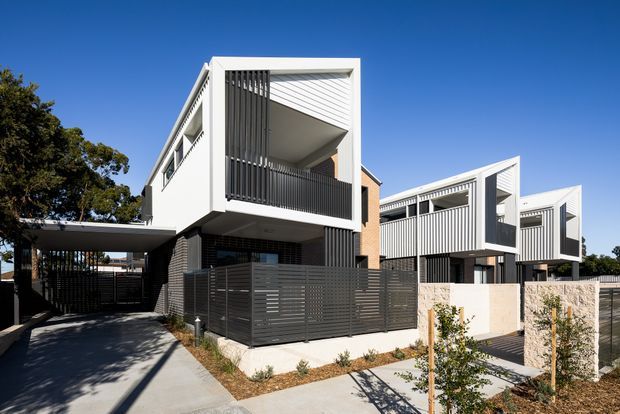 Street-level exterior view of a contemporary multi-unit residential development with three-storey forms, crisp white rendered façades, dark vertical screening and balcony balustrades, recessed upper-level terraces, brick side walls, and gated private front courtyards. A covered carport and driveway are visible at left, with young landscaping along the pedestrian path.