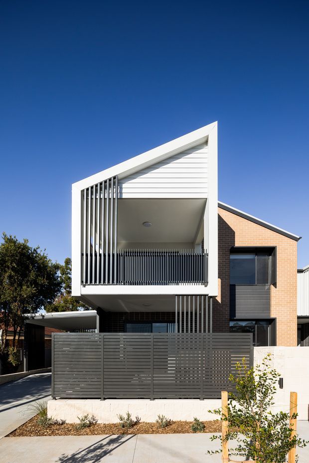 Street-facing view of a contemporary multi-storey townhouse or residential building with a strongly geometric gable-like facade. The facade combines warm red brick, white horizontal cladding, dark grey horizontal privacy fencing, black vertical balcony screens, and projecting balconies. A steep triangular roofline and asymmetrical upper-level balcony create a sculptural profile, with a paved driveway and small front garden in the foreground under a clear blue sky.