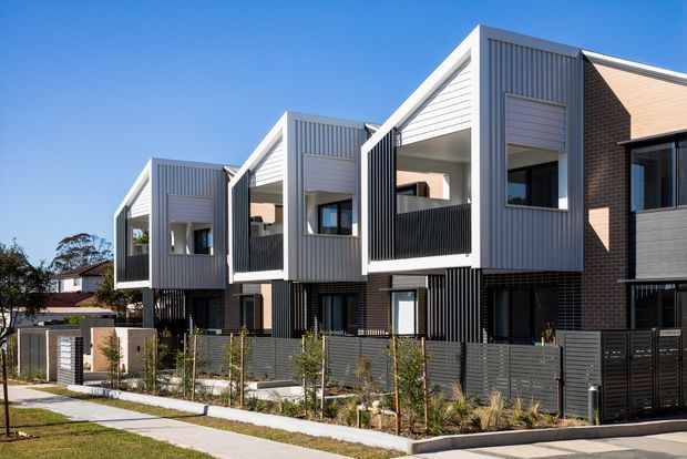 Contemporary multi-unit residential development with a repeating series of projecting gabled forms, light grey vertical metal cladding, dark brick and lower-level privacy screens. The facade includes recessed balconies or terraces, large windows, landscaped front setbacks, and shared pedestrian access under a clear blue sky.