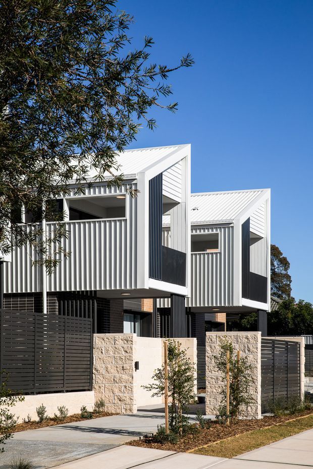 Street-facing exterior view of a contemporary multi-storey residential development with repeated angular gabled forms, white corrugated metal cladding and roofing, dark vertical screening, projecting upper-level volumes, and masonry boundary walls. The property includes landscaped front setbacks, private fencing, and pedestrian access.