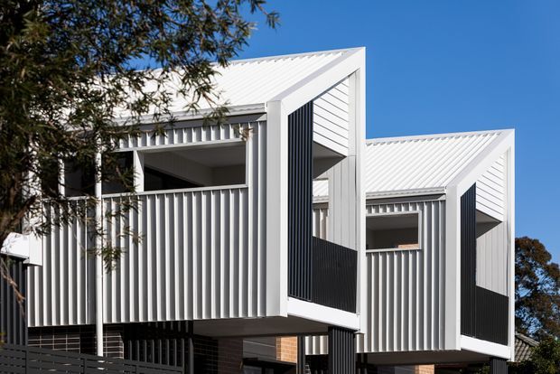 Exterior architectural photograph of a contemporary multi-level residential building with repeated angular gabled forms, white corrugated metal roofing and cladding, vertical ribbed screens, dark recessed openings, and projecting balcony-like volumes. Trees frame the edges against a clear blue sky.