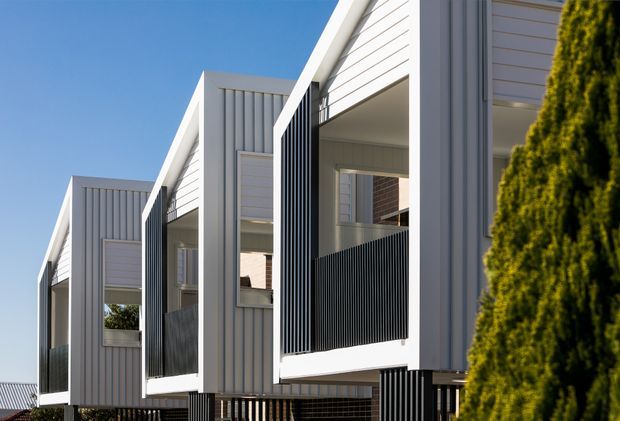 Exterior architectural photograph of a contemporary row of compact multi-storey residences with repeating gabled forms, pale vertical cladding, white structural frames, recessed balconies, and black vertical privacy screens. A tall evergreen hedge partially frames the right side, with a clear blue sky behind.