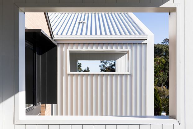 A close-up architectural view through a large rectangular opening or framed portal, showing a small cream corrugated-clad structure with a matching corrugated pitched roof, a projecting rectangular window surround, and dark-framed glazing at left. The scene is brightly lit with clean contemporary detailing and strong geometric lines.