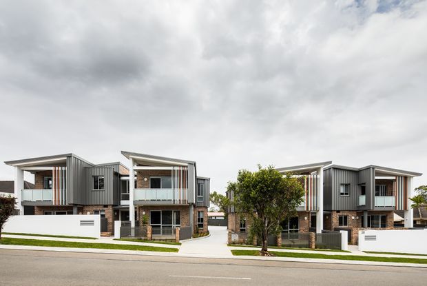 Street-facing view of a contemporary multi-unit residential development composed of several attached or closely grouped two-storey townhouses. The buildings feature flat and shallow skillion roof forms, grey vertical metal cladding, exposed brick walls, white rendered boundary walls, balconies, and landscaped front yards.