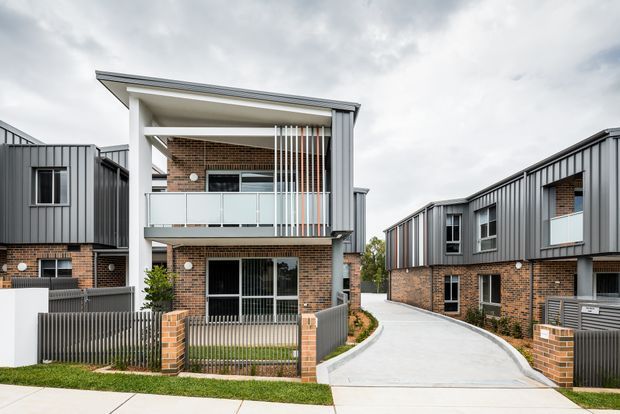 Exterior view of a contemporary multi-unit residential development with two-storey brick townhouses, dark vertical metal cladding, projecting balconies, white structural frames, and metal privacy screens. A paved driveway curves between the buildings, with small fenced front gardens and brick gate pillars under an overcast sky.