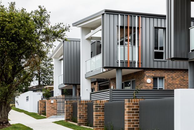 Exterior streetscape view of a contemporary multi-storey residential development, featuring dark vertical metal cladding, exposed brickwork, white rendered structural elements, glass balcony balustrades, and integrated private front courtyards with metal gates and brick piers.