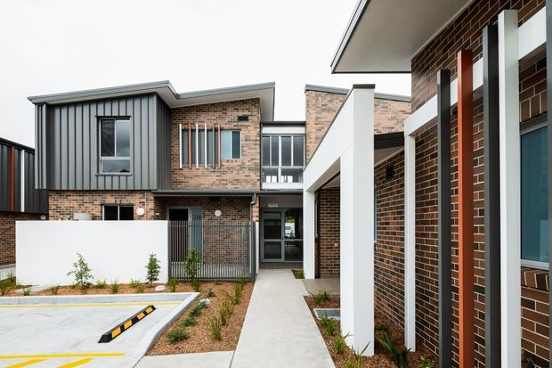 Exterior view of a contemporary multi-unit brick building with a central pedestrian walkway, landscaped courtyard planting, covered entry structure, dark metal cladding, and contrasting white architectural columns and screens. The complex features flat and low-pitched roof forms, varied massing, and vertical window detailing.