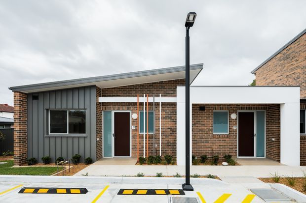 Street-facing view of a contemporary low-rise brick residential development with several compact attached units. The facade combines exposed brown brick, dark vertical metal cladding, large rectangular windows, recessed entries, flat or very low-pitched roof planes, and simple landscaped front strips. A paved driveway or shared parking area with marked bays and speed humps occupies the foreground.
