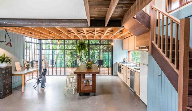 A bright open-plan industrial-style interior combining a kitchen, dining/work area, and compact stairway. The room features expansive black-framed glazing, exposed timber roof trusses and beams, a polished concrete floor, pale blue vertical wall panelling, warm timber cabinetry and staircase joinery, and a central timber island or worktable. A loft-like timber structure is visible overhead, with greenery seen through the glazing.