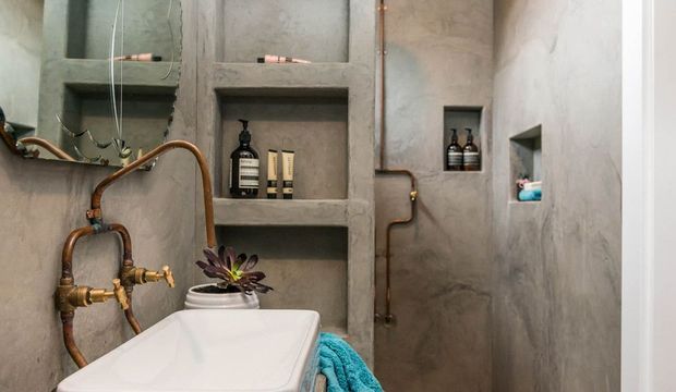 Compact industrial-style bathroom featuring a white freestanding bathtub, exposed aged copper plumbing and brass fittings, and built-in recessed shelving niches in textured grey plaster walls. Toiletries and a small plant occupy the niches, with a turquoise towel adding a restrained colour accent.