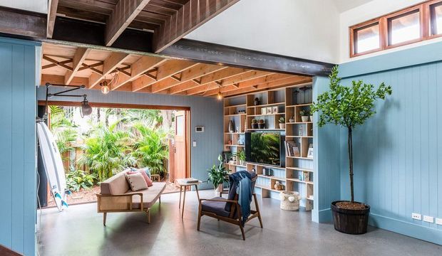 Bright, open-plan living room with a mid-century-inspired seating arrangement, polished concrete floor, blue vertical wall panelling, extensive built-in shelving, exposed timber roof framing, and a large opening to lush outdoor greenery. High clerestory windows and exposed structural beams create an airy, characterful interior.