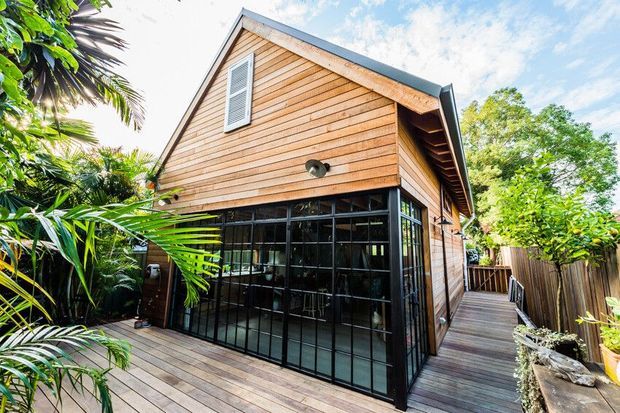 Exterior view of a compact timber-clad gable-roofed building surrounded by lush tropical planting. The structure has warm horizontal timber weatherboard cladding, dark metal-framed floor-to-ceiling glazing with black grid divisions, a timber deck and walkway, and a small high-level louvered window. The image emphasizes the building facade and outdoor deck setting.