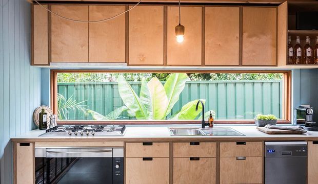 A compact, contemporary kitchen with warm timber cabinetry, pale blue vertical wall panelling, and a long horizontal window overlooking lush greenery and a green fence. The kitchen includes a gas cooktop and oven, stainless-steel sink and dishwasher, open shelving with bottles, and a simple pendant light. The overall palette combines natural wood, soft blue, white, and stainless steel in a bright, functional space.