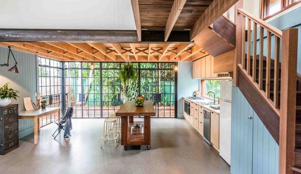 A bright open-plan kitchen and work area beneath a dramatic exposed timber roof structure. The room features polished concrete flooring, pale blue vertical wall panelling, extensive black-framed industrial-style glazing, timber cabinetry, a central wooden island with stools, a desk area, and a prominent timber staircase leading to an upper level. Hanging plants and greenery are visible beyond the glazing.