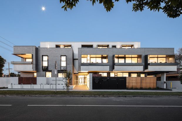 Photograph of a contemporary multi-storey residential apartment or townhouse development viewed from across a street at dusk. The facade combines dark grey masonry or concrete-like cladding, extensive warm-lit glazing, projecting balconies, vertical timber screening, black garage or entry doors, and a light ribbed upper roof-level volume. A low front wall, driveway access, street, and sparse landscaping are visible.