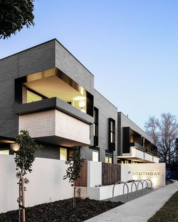 Exterior architectural photograph of a contemporary multi-unit residential development with a long, linear form and flat roof. The building is finished in dark grey brick with projecting white timber-clad balcony volumes, black metal balcony balustrades and deep recessed openings. A landscaped frontage includes a white rendered entry wall with timber screening, signage reading “SOUTH BAY,” young trees, and bicycle hoops along the footpath.