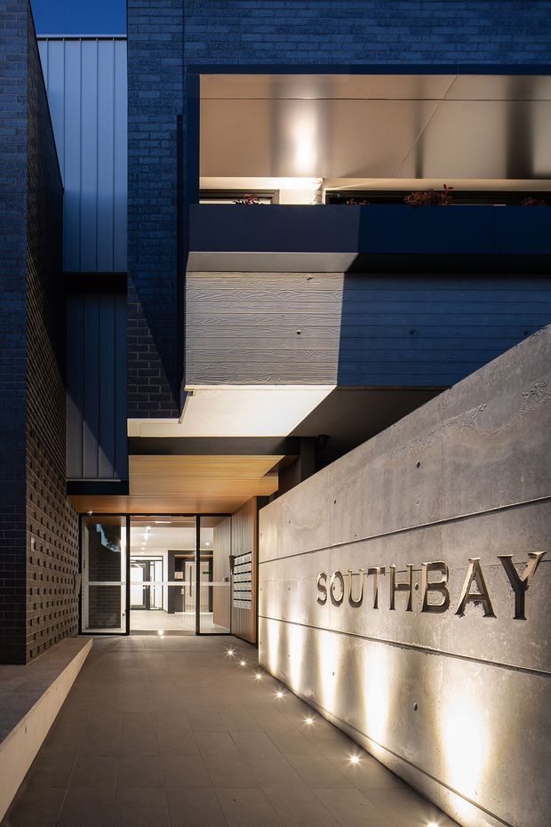 A vertical architectural photograph of a contemporary multi-storey building entrance. The recessed entry is approached along a paved walkway between dark brick walls and a large raw concrete wall bearing the raised word “SOUTHBAY.” Warm concealed and ground-level lighting illuminates the entrance, timber soffit, concrete surfaces, dark metal-framed glazing, and projecting upper balconies. The composition emphasises the building facade and entry sequence.