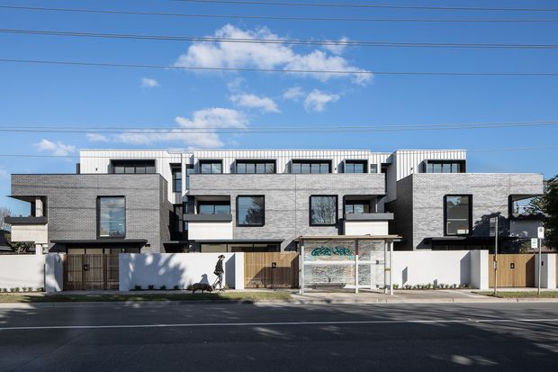 Street-facing view of a contemporary multi-unit residential development with several attached or closely grouped homes. The building has a strongly rectilinear composition, grey brick masonry façades, white upper-level cladding, dark-framed windows, projecting box-like balconies, and private front courtyards screened by white walls and timber gates. A broad paved street and overhead utility wires are visible in the foreground and sky.