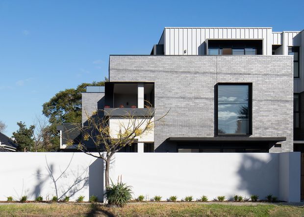 Exterior photograph of a contemporary multi-storey residence with a rectilinear composition, light grey brick facade, white vertical metal cladding at the upper level, dark-framed windows, recessed balconies, and a tall white boundary wall with simple landscaping. The home has a flat or very low-pitched roof and a clean, minimalist facade under a clear blue sky.
