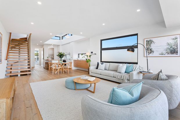 Bright open-plan contemporary living and dining area with pale timber flooring, white walls and ceiling, a large light-grey sectional sofa, curved armchairs, a round timber coffee table, and pale blue accent cushions. A timber open-riser staircase and dining table sit to the left, while a large black-framed window, skylights, recessed lighting, and coastal artwork add architectural and decorative interest.