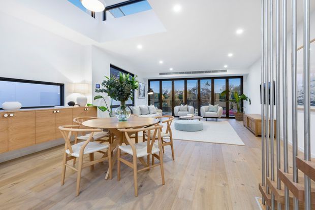 Bright open-plan living and dining area with light timber flooring, a round timber dining table and chairs in the foreground, and a lounge seating zone beyond. The room features white walls and ceiling, recessed lighting, expansive black-framed glazing, a skylight, built-in timber media cabinetry, indoor plants, and a vertical timber screening element at the right.