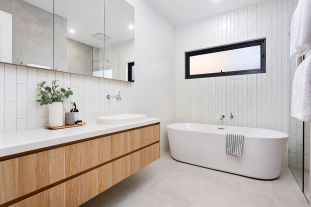 Bright contemporary bathroom featuring a freestanding white bathtub, floating timber-veneer vanity with integrated basins, white vertical rectangular wall tiles, pale floor tiles, large mirror, and a black-framed horizontal window. The room has a clean, calm, minimalist appearance with natural timber accents.