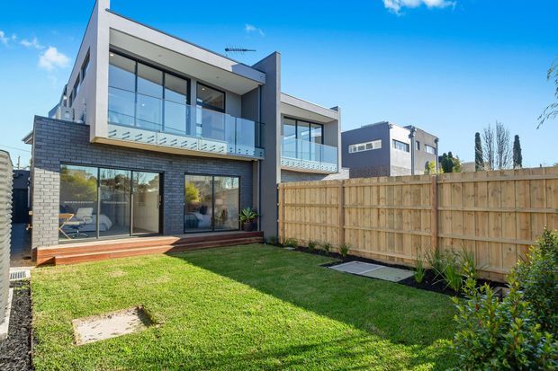 Rear exterior view of a contemporary multi-storey townhouse with dark brick lower-level cladding, white rendered upper walls, extensive glazing, glass balcony balustrades, and a small private lawn enclosed by timber fencing.