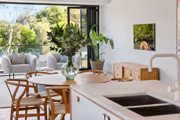 Bright open-plan interior combining a contemporary kitchen and dining area with a living space beyond. A light timber dining table and curved wooden chairs sit beside a white kitchen island with a dark double-basin sink. Large glazed doors open to leafy outdoor views, while a sculptural floral arrangement, potted plant, timber sideboard and artwork add warmth.