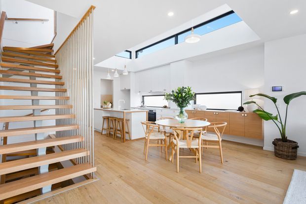 Bright open-plan kitchen and dining area with a minimalist white fit-out, warm timber flooring, light timber cabinetry, a round dining table with timber chairs, and a floating open-riser staircase. A skylight and high-level glazing bring in abundant natural light, while vertical timber balusters define the stair edge.