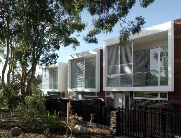 Contemporary multi-level residential facade with crisp white projecting box forms, extensive glazing, horizontal external louvres, and dark timber-toned cladding accents. Black vertical fencing and dark brickwork define the lower level, while mature eucalyptus trees and landscaped planting frame the building.