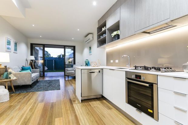 Bright open-plan kitchen and living area with white cabinetry, grey wood-look overhead cupboards, a pale stone benchtop, stainless-steel appliances, and a neutral splashback. Warm timber flooring extends into the adjacent living room, which includes a sofa, armchair and coffee table. Glazed sliding doors open to a small outdoor area, and recessed ceiling lights provide even illumination.
