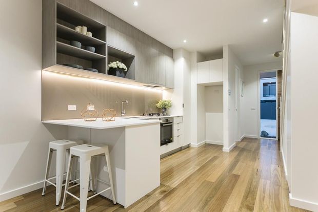 Bright contemporary galley kitchen opening into a hallway, with a compact white island and two stools, integrated cabinetry, open shelving, under-cabinet lighting, stainless tap and appliances, and warm timber flooring. The space has a clean, refined apartment-style fit-out with recessed ceiling lights.
