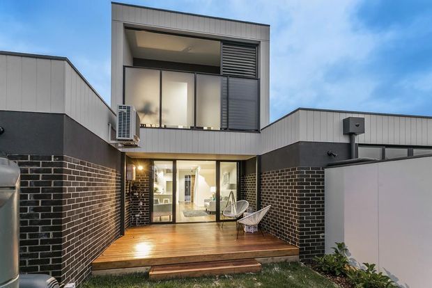 Rear exterior view of a contemporary multi-level residence with a dark brick lower facade, light vertical metal cladding above, broad glazed doors and windows, and a timber outdoor deck opening from the living area. The enclosed courtyard-like setting includes outdoor seating and a rooftop or upper-level balcony zone with louvred screening.