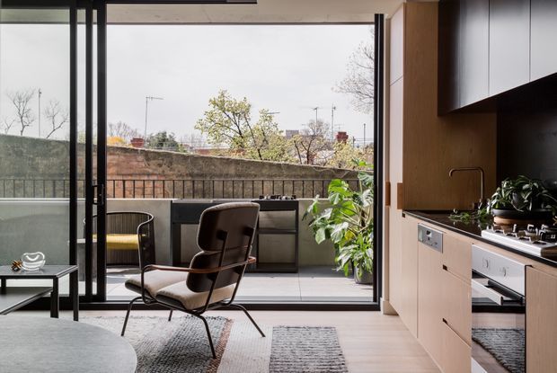 A contemporary open-plan living and kitchen area opening onto a balcony through large black-framed sliding glass doors. The room has light timber flooring, pale timber kitchen cabinetry, a dark benchtop and backsplash, integrated appliances, and a modern lounge chair with a small side table. Outdoor balcony seating and planted greenery are visible beyond the glazing, with an urban residential outlook.
