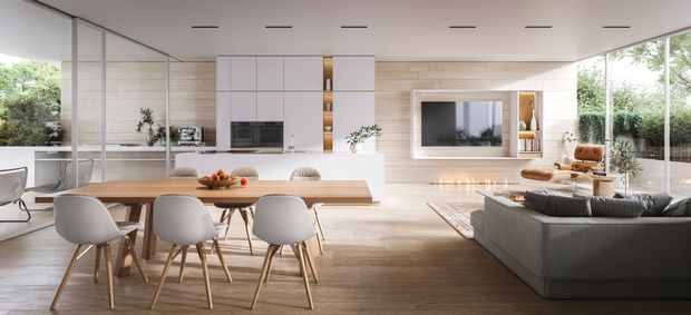 A bright, open-plan contemporary living, dining and kitchen area with floor-to-ceiling glazing on two sides. The space features a streamlined white kitchen with integrated appliances and timber-look cabinetry or wall panelling, a long dining table with modern upholstered chairs, and a large grey sectional sofa facing a wall-mounted television and built-in shelving. Warm timber flooring, pale finishes, recessed linear ceiling lights, and abundant greenery visible outside create an airy, refined atmosphere.