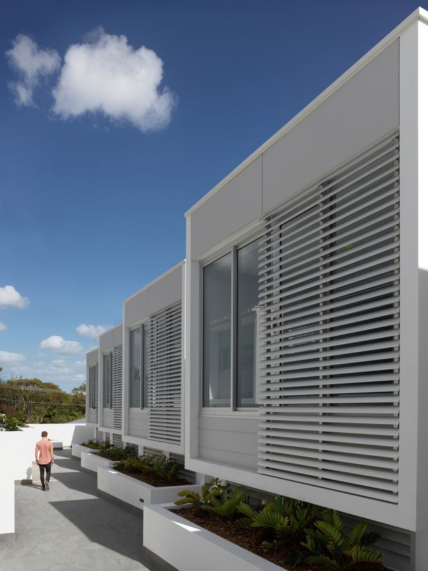 Exterior view of a long, modern white building with a sharply sloping roofline, large glazed openings, and extensive horizontal louvred sun-shading screens. White raised planter boxes with low tropical planting line the paved walkway, while a person provides scale. The image is photographed under a bright blue sky.
