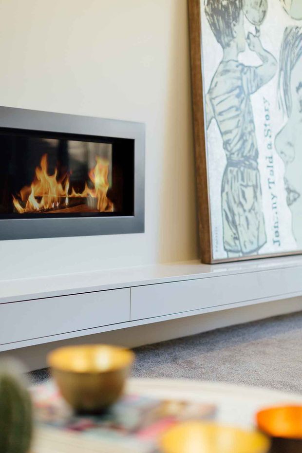 Close-up interior view of a contemporary living room media/fireplace wall. A built-in linear gas fireplace with a dark metal surround is set into a pale wall above sleek white handleless cabinetry. A large framed artwork leans on the joinery, with a soft grey carpet and blurred coffee-table objects in the foreground.