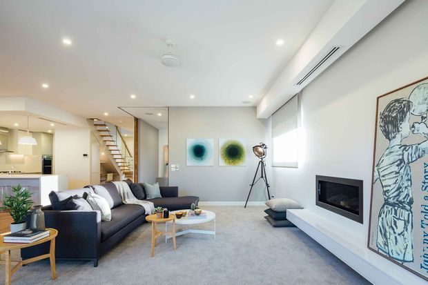 Bright open-plan contemporary living room with a large grey sectional sofa, light carpet, low timber-and-white coffee table, wall-mounted fireplace, recessed lighting, and a minimalist media wall. The kitchen and dining area are visible in the background, separated by a stairway with open timber treads and a slim metal balustrade. Neutral white and grey finishes create an airy, refined interior.