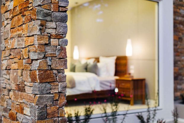 Close-up exterior view of a rustic stacked-stone wall beside a large window, with a softly blurred bedroom visible through the glazing. The stonework combines warm brown, rust and grey tones, while the bedroom contains a bed, bedside table and warm lighting.
