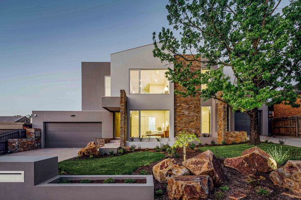 Contemporary two-storey residential facade viewed from the street, featuring rectilinear rendered volumes, extensive glazing, a prominent natural stone-clad vertical feature, an integrated double garage, and a landscaped front garden with large decorative rocks and low retaining walls.
