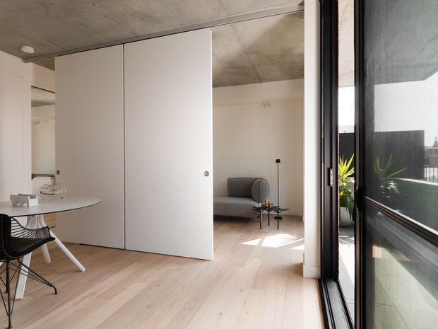 Bright, minimalist open-plan apartment interior with pale timber flooring, white walls and full-height black-framed glazing. A compact dining area sits in the foreground, while a grey sofa occupies the living area beyond large white flush-panel doors. The exposed concrete ceiling and restrained furnishings create a clean contemporary atmosphere, with a balcony visible through the glazing.