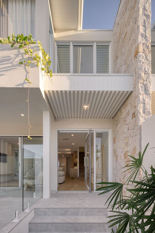 A bright contemporary residential exterior viewed from a raised terrace or entry approach. The facade combines white rendered walls, a pale irregular stone-clad wall, a projecting upper balcony with a ribbed soffit, louvred windows, and large glazed sliding doors opening into a warm-toned interior. Pale stone steps, glass balustrading, and tropical-style planting frame the entrance.