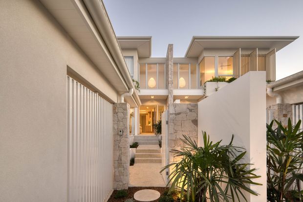 Contemporary multi-level residential facade viewed from a narrow landscaped entry walkway. The home features pale rendered walls, broad flat roof overhangs, warm illuminated glazing, vertical privacy screens, and prominent natural stone-clad columns and walls. A gated garage or entry structure lines the left side, while tropical-style planting and stepping surfaces lead toward the recessed front entrance.