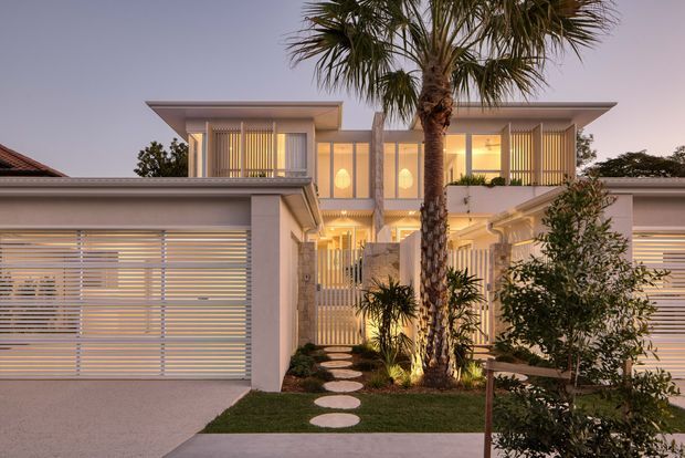 Twilight exterior view of a contemporary two-storey residence with a symmetrical composition, flat projecting rooflines, pale rendered walls, extensive vertical screening, illuminated glazing, and integrated garage doors. A landscaped entry path with circular stepping stones, lawn, palms, and shrubs leads toward the central entrance.