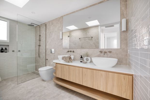Bright contemporary bathroom with a walk-in glass shower, wall-mounted toilet, freestanding vessel basin, and a long floating timber vanity. Large mirrors, skylights, pale stone-look surfaces, and light grey tiled walls create an airy, refined space.