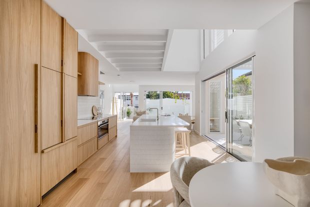 Bright, open-plan contemporary kitchen and dining area with a long light-wood wall of integrated cabinetry, a white tiled island with seating, pale timber flooring, and large sliding glass doors opening to an outdoor area. A white dining table occupies the foreground, while strong daylight enters through the glazing and a high-level opening.