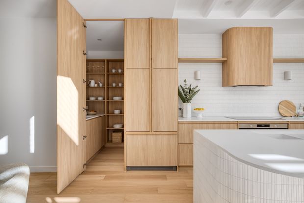 Bright, refined contemporary kitchen with extensive light timber cabinetry, a concealed walk-in pantry, white tiled splashback, integrated appliances, and a curved island clad in small white tiles. The room has a light timber floor, white walls, and a distinctive ribbed ceiling with exposed beams. Sunlight creates strong geometric highlights across the cabinetry and island.