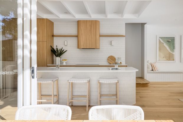 Bright open-plan kitchen and living area viewed from a dining space. The kitchen features a curved white tiled island with three woven bar stools, light timber cabinetry, white tiled splashback, open timber shelving, and a white coffered ceiling. Light timber flooring continues into the adjacent seating area, which includes a built-in bench and framed coastal artwork. Full-height glazing and white-framed doors bring in abundant natural light.